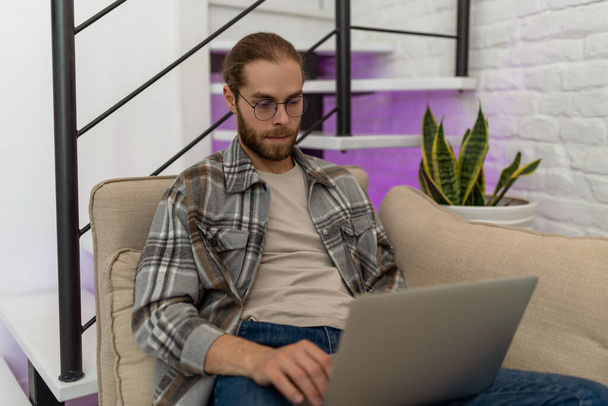 Young attractive serious concentrated guy in glasses browsing at his laptop, sitting on sofa at home, wearing casual outfit. - Фото, зображення