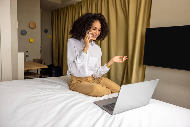 En el hotel. Mujer de negocios de pelo rizado hablando por teléfono en la habitación de hotel - Foto, Imagen