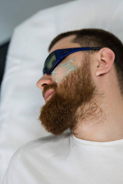 Bonito homem está deitado em uma cama no escritório cosmetologists, prepara-o para um procedimento de depilação a laser na barba. Esteticista aplicou gel em seu rosto em uma clínica de beleza - Foto, Imagem