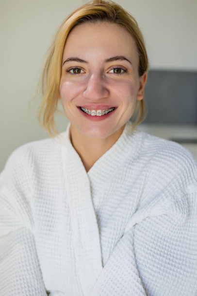 Positive girl with natural beauty face with out make up, wearing braces and a white robe, smiles at the camera after beauty procedures, skin rejuvenation, in a beauty clinic, spa salon - Photo, Image