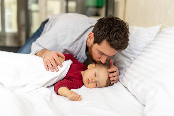 Brunette bearded father kissing sleeping infant baby tenderly home interior. - Фото, изображение
