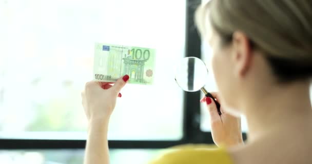 Kobieta przegląda szkło powiększające za 100 euro i sprawdza autentyczność filmu 4k w zwolnionym tempie. Koncepcja fałszywych pieniędzy - Materiał filmowy, wideo