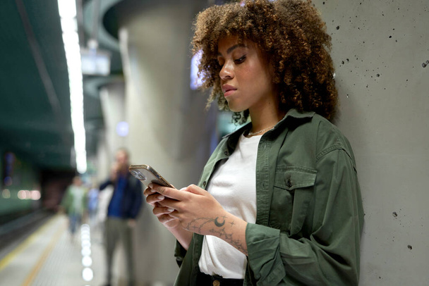 Młoda afro kobieta przegląda telefon komórkowy i czeka na metro  - Zdjęcie, obraz