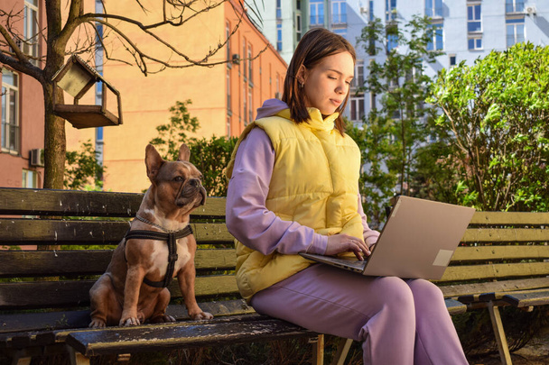 Beautiful young female freelancer businesswoman using laptop while sitting on bench with her adorable small dog at urban area in sunny day. City lifestyle concept, remote work. Woman working outdoors - Photo, Image