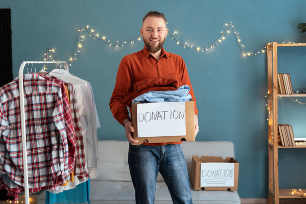 Η ιδέα της δωρεάς. Άντρας που κρατάει ένα κουτί δωρεών με γεμάτα ρούχα κοιτάζοντας την κάμερα. Αντιγραφή χώρου - Φωτογραφία, εικόνα