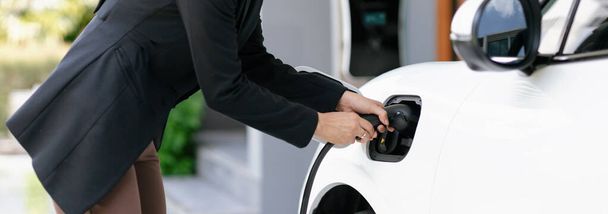 Closeup progressive woman install cable plug to her electric car with home charging station. Concept of the use of electric vehicles in a progressive lifestyle contributes to clean environment. - Φωτογραφία, εικόνα
