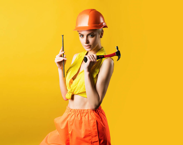 Sexy woman in helmet to use hammer. Young woman hammering nail at workshop on yellow background isolated. Female model working on building. Everything can be fixed concept - Foto, Imagem