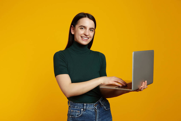 Portrait of a smiling young brunette girl holding laptop computer isolated over yellow background. - Фото, изображение