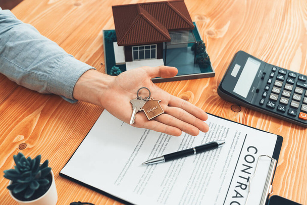 Real estate agent presents house key on his hand over wooden table surrounded by calculator and legal documents, as a symbolic gesture for persuasive home sales and mortgage offer. Entity - Foto, Imagem