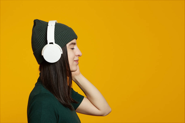 Seitenansicht der jungen Frau in grünem Outfit, Musik hören in weißen Kopfhörern mit geschlossenen Augen, isoliert auf gelbem Hintergrund mit leerem Platz für Text - Foto, Bild