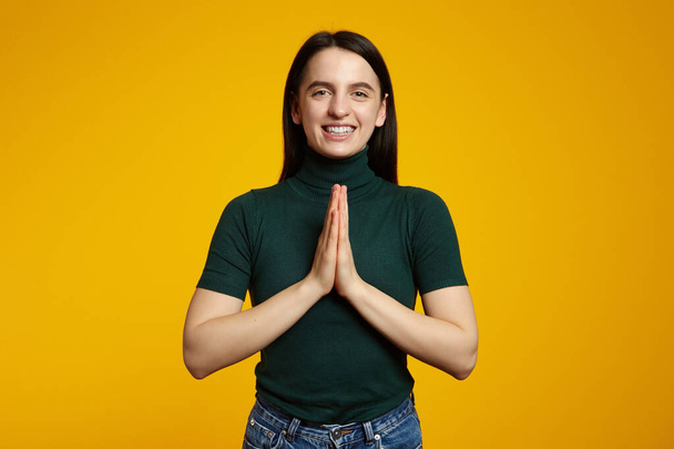Hopeful young girl keeps hands together in praying gesture, enjoys peaceful atmosphere, has glad expression, isolated over yellow background - Foto, afbeelding