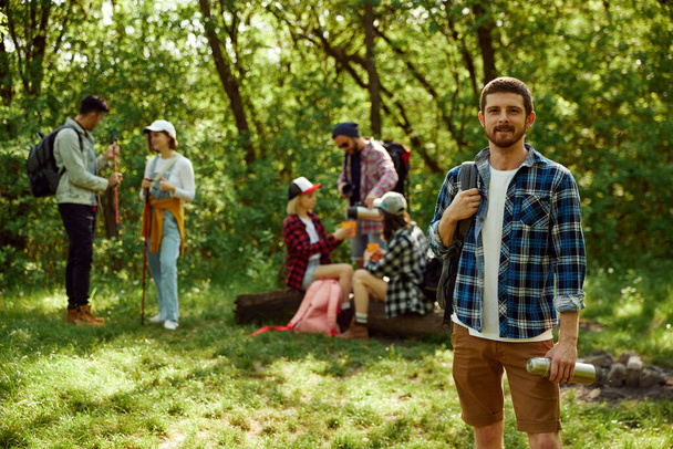 Young happy, smiling man in checkered shirt going hiking with friends. Group of young people walking in forest. Concept of active lifestyle, nature, sport and hobby, friendship, leisure time - Photo, image