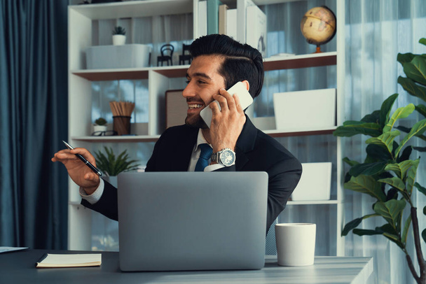 Трудолюбивый бизнесмен занят разговорами по телефону с клиентами, работая с ноутбуком в своем офисе, как концепция современного трудолюбивого офисного образа жизни с мобильным телефоном. Пылкий - Фото, изображение