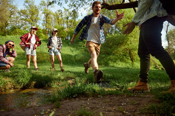 Grupo de amigos, jovens ativos caminhando, andando na floresta, pulando sobre o rio e se divertindo ao ar livre no dia quente da primavera. Conceito de estilo de vida ativo, natureza, esporte e hobby, amizade - Foto, Imagem