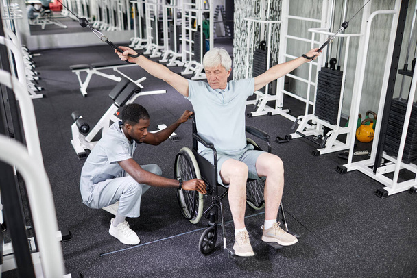 Porträt einer afroamerikanischen Rehabilitationstherapeutin, die einem älteren Mann im Rollstuhl bei körperlichen Übungen im Fitnessstudio hilft - Foto, Bild