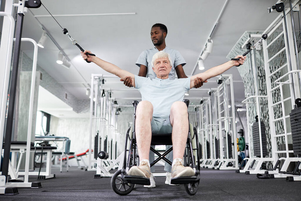 Πορτραίτο χαμηλής γωνίας του Αφροαμερικανού θεραπευτή αποκατάστασης που βοηθά τον ηλικιωμένο σε αναπηρική καρέκλα να κάνει σωματικές ασκήσεις στο γυμναστήριο - Φωτογραφία, εικόνα
