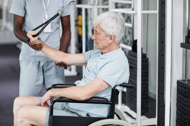 Portrait latéral d'un aîné en fauteuil roulant faisant des exercices de réadaptation au gymnase à la clinique, espace de copie - Photo, image