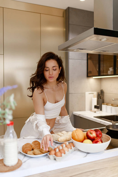 Великолепная сексуальная молодая темноволосая женщина, опираясь на кухонный стол, тянется за яйцом. Концепция питания и приготовления пищи - Фото, изображение