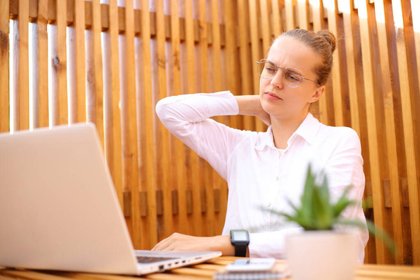 Exhausted woman wearing white shirt sitting in outdoor cafe working on laptop massaging her painful neck frowning face looking at computer display. - Foto, Bild
