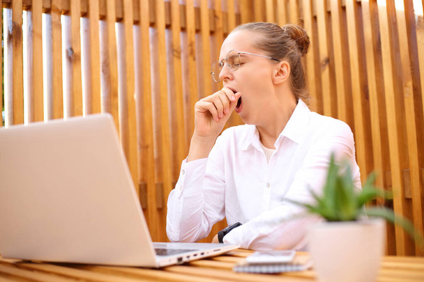 Sleepy overworked woman wearing white shirt sitting in outdoor cafe working on laptop yawning needs rest covering mouth with fist doing online project. - Foto, Imagen