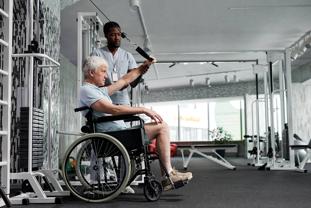 Tekerlekli sandalyeyi spor salonunda kullanan ve asistanıyla rehabilitasyon egzersizleri yapan yaşlı bir adamın yan görünümü. - Fotoğraf, Görsel