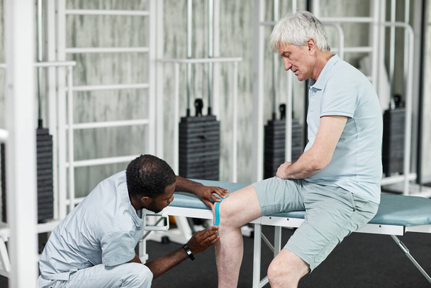 Porträt des Rehabilitationsspezialisten beim Tapen am Knie eines älteren Patienten in der Physiotherapie-Klinik, Kopierraum - Foto, Bild