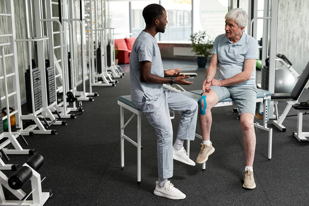 Full length portret van revalidatie therapeut consulting senior patiënt in de sportschool in kliniek, kopieerruimte - Foto, afbeelding