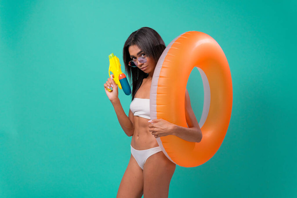 african woman dancing with an inflatable circle in a swimsuit with a water gun in her hands, isolated over blue background. - Фото, зображення