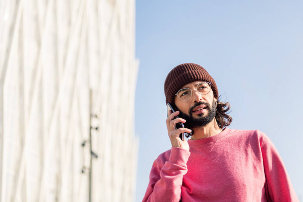 stylish bearded young man in pink jersey talking on smart phone, concept of modern lifestyle and technology of communication, copy space for text - Photo, Image