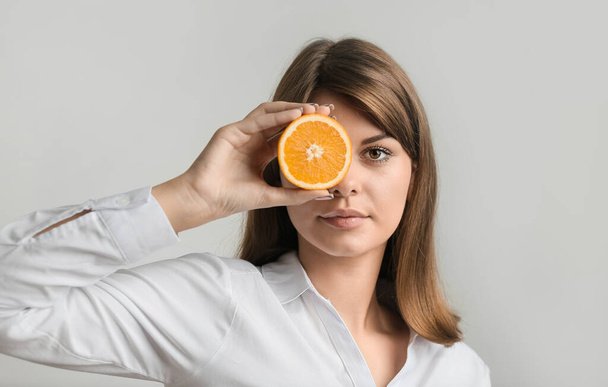 Dziewczyna z pomarańczą reklamuje krem. Zdrowa skóra. Dziewczyna w białej koszuli zamyka oko pomarańczą na szarym tle - Zdjęcie, obraz
