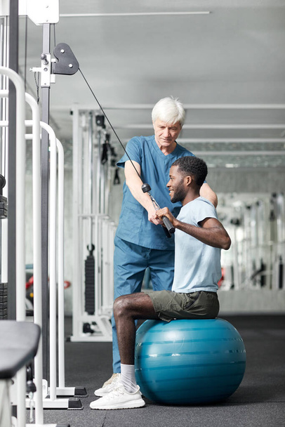 Full length portret van fysiotherapie specialist helpen Afro-Amerikaanse jongeman doen revalidatie oefeningen in de sportschool, kopiëren ruimte - Foto, afbeelding