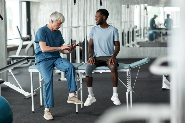 Retrato completo del terapeuta de rehabilitación senior consultando a un joven negro en la clínica - Foto, Imagen