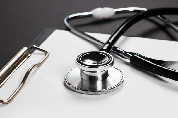 A clipboard and stethoscope with white paper on it, set against a dark background. - Foto, Imagen