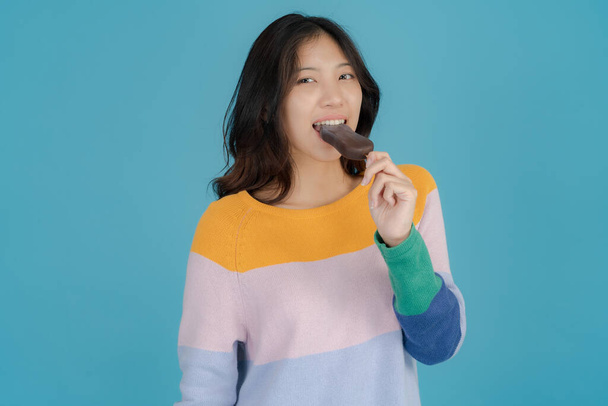 Gyönyörű fiatal nő mosolyog boldogan, miközben tartja és eszik fagylaltot a kezében a kék háttér - Fotó, kép