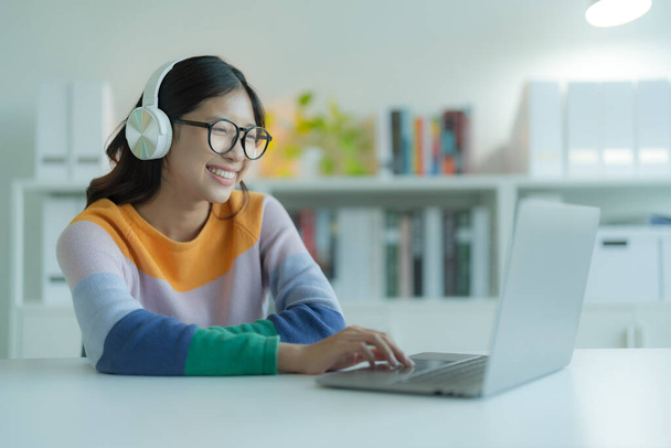 Egy fiatal nő vagy diák laptopot használ a könyvtárban, miközben szemüveget és fejhallgatót visel. Mosolyog és boldognak tűnik.. - Fotó, kép