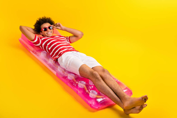 Plná délka fotografie cool funky chlap nosit pruhované tričko s odpočinkem nafukovací postel prázdný prostor izolované žluté barvy pozadí. - Fotografie, Obrázek