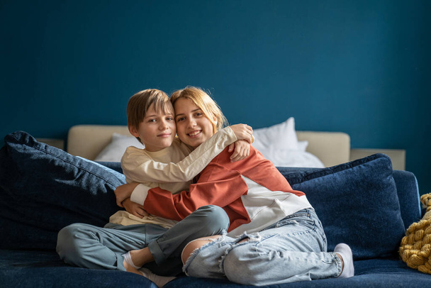 Sibling friendship. Portrait of happy children brother and sister hugging embracing, enjoying playing and spending leisure time together at home. Teen girl and her younger brother resting on sofa - Photo, Image