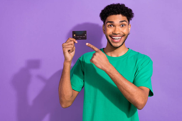 Photo of astonished handsome guy indicate finger debit card wear green t-shirt isolated on violet color background. - Fotografie, Obrázek