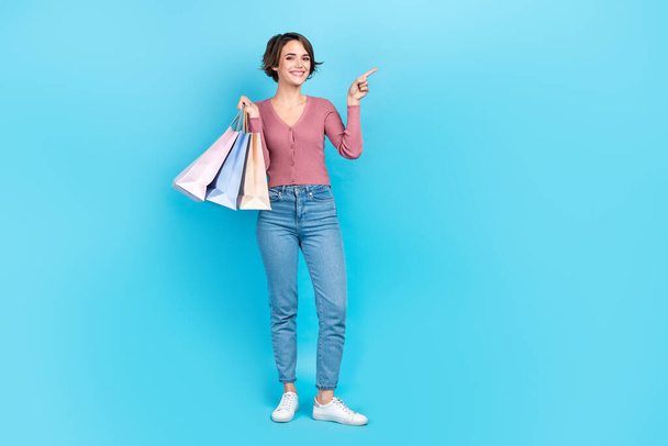 Full length photo of pretty cheerful person hold boutique bags direct finger empty space isolated on blue color background. - Фото, изображение