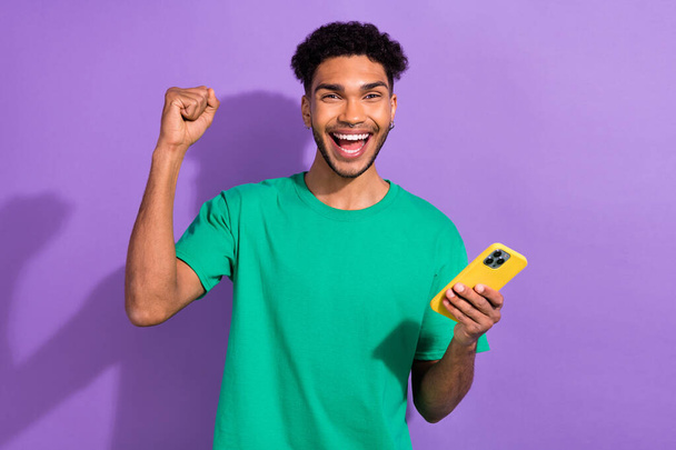 Photo of cheerful lucky man wear green t-shirt winning game apple samsung iphone modern gadget isolated purple color background. - Fotó, kép