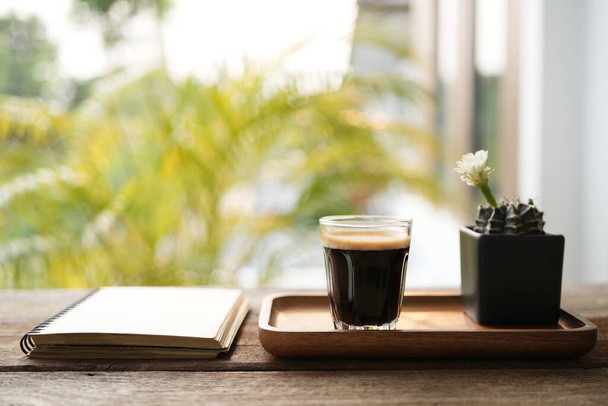 taza de vidrio de café y flor de cactus blanco en una olla negra en la mesa de madera con cuaderno - Foto, Imagen