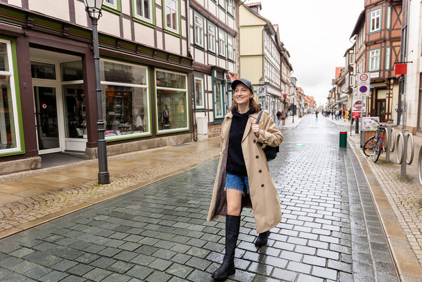 Fiatal felnőtt divatos elegáns gyönyörű kaukázusi boldog mosolygós nő élvezze séta gyalogos terület Európa esős nap. Női személy portré viselni farmer nadrág biege ballon kabát városi utcában. - Fotó, kép