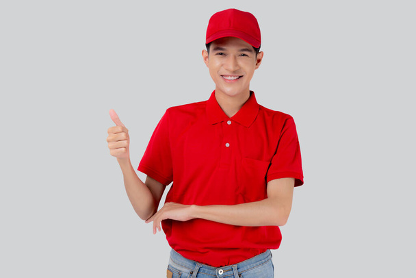 Joven hombre asiático en uniforme rojo y gorra de pie pulgares hacia arriba con confianza aislado sobre fondo blanco, empleado o distribuidor, mensajero y entrega, repartidor y expresión, logística y carga. - Foto, Imagen