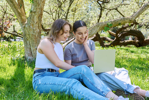 Two teen girls college high school students sitting in park on grass looking at laptop. Technologies for leisure communication, study, internet online services, e-shopping - 写真・画像