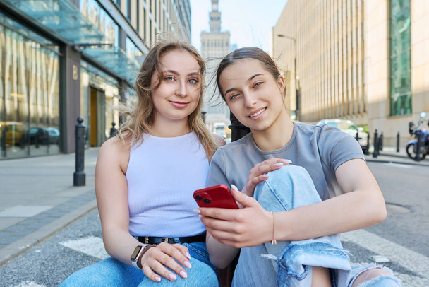 Two smiling teenage female students with smartphone, modern urban style buildings road background. Fashionable girls sitting on sidewalk. Youth fashion style city lifestyle, online internet technology - Fotoğraf, Görsel