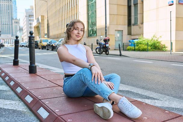 Young beautiful fashionable girl 19, 20 years old sitting on sidewalk, modern urban style. Natural blonde woman with blue eyes, curly hair looking at camera, european city, road background - Fotografie, Obrázek