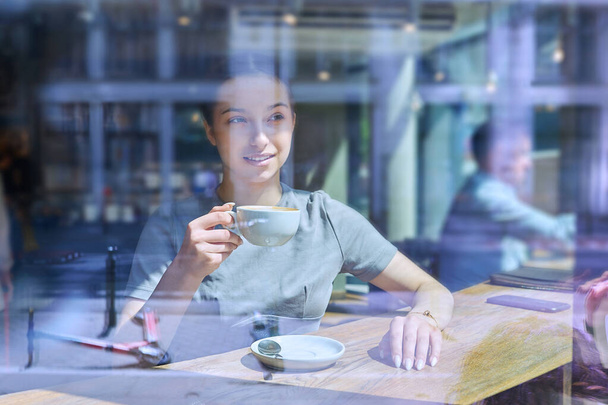 Beautiful young female inside cafe with cup of coffee, view through glass window. Attractive teenage girl resting in cafe. Beauty, youth, lifestyle concept - Foto, afbeelding