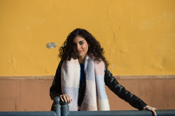 pretty young woman with curly brunette hair against a yellow background is dressed in winter clothes and wearing a scarf to protect herself from the cold. The woman is happy and having fun. - Foto, imagen