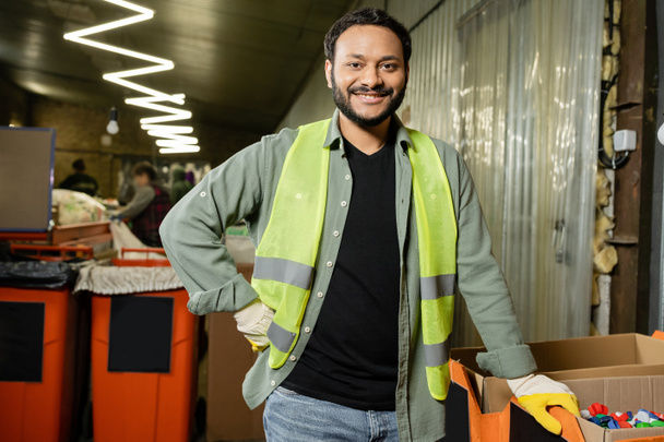Smiling indian worker in high visibility vest and protective gloves looking at camera near plastic caps in carton boxes while working in waste disposal station, garbage sorting and recycling concept - Photo, Image