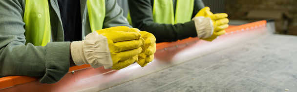 Cropped view of sorter in protective gloves and vest standing near conveyor and blurred colleague while working in garbage sorting center, garbage sorting and recycling concept, banner  - Photo, Image